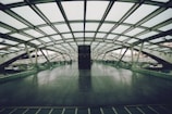 A spacious, modern pedestrian bridge with a glass roof and floor. The structure features a grid-like pattern and is mostly empty, with a couple of people in the distance. The bridge is surrounded by urban elements like cars and buildings, seen through the transparent sides.