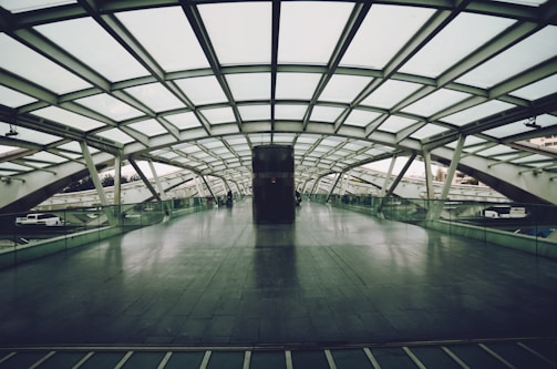 A spacious, modern pedestrian bridge with a glass roof and floor. The structure features a grid-like pattern and is mostly empty, with a couple of people in the distance. The bridge is surrounded by urban elements like cars and buildings, seen through the transparent sides.