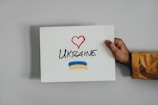 A hand holds a white sign with a red heart drawn above the word 'Ukraine' written in black. Below the text, there are blue and yellow brush strokes resembling the Ukrainian flag. The hand is wearing a brown or tan sleeve.