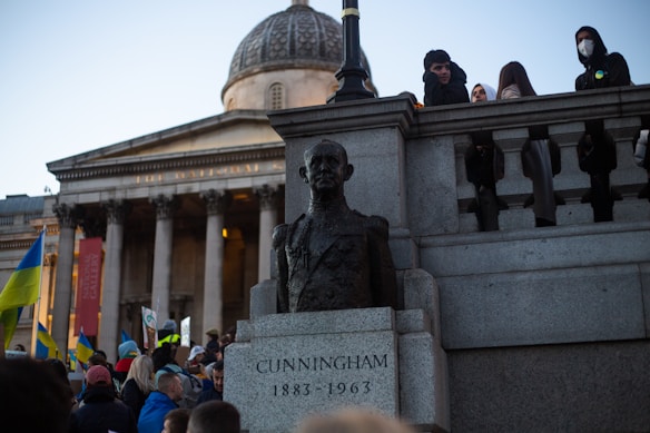 A public square with a large classical-style building featuring columns and a dome in the background. A stone bust of Cunningham is prominently displayed in the foreground. People are gathered around, some holding flags with a yellow and blue color scheme, suggesting a demonstration or public event.