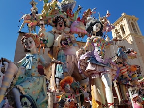 Large and colorful sculptures featuring exaggerated and whimsical characters in elaborate costumes. The scene includes various figures with vibrant outfits and intricate details, set in an outdoor environment with a blue sky in the background.