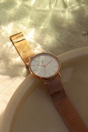 A gold mesh strap wristwatch with a white analog dial is placed on a smooth, cream-colored surface. Soft, dappled sunlight creates gentle patterns and highlights on the surface, emphasizing the elegant design of the watch.