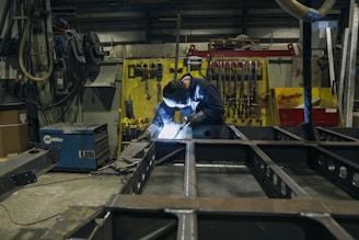 A person is engaged in welding work inside an industrial workshop. The environment is cluttered with various tools and equipment, including a welding machine. Bright light from the welding process illuminates the workspace. The walls are lined with hanging tools and yellow and red equipment in the background.