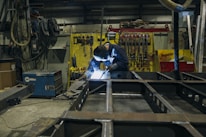 A person is engaged in welding work inside an industrial workshop. The environment is cluttered with various tools and equipment, including a welding machine. Bright light from the welding process illuminates the workspace. The walls are lined with hanging tools and yellow and red equipment in the background.