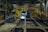 A person is engaged in welding work inside an industrial workshop. The environment is cluttered with various tools and equipment, including a welding machine. Bright light from the welding process illuminates the workspace. The walls are lined with hanging tools and yellow and red equipment in the background.
