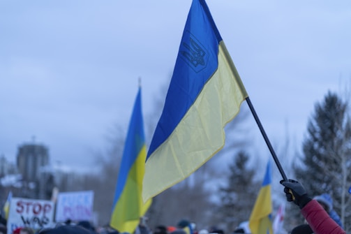 A large Ukrainian flag is held aloft amidst a protest or rally, with several other similar flags visible in the background. Signs with messages are held by participants, and the scene takes place outdoors on a cloudy day with trees and a building in the distance.