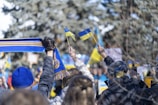 A group of people in winter clothing holding small blue and yellow flags are gathered together. One person is holding a blue and yellow scarf, and several others are taking pictures with their phones. The background shows a wintry outdoor setting with blurred trees.