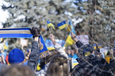 A group of people in winter clothing holding small blue and yellow flags are gathered together. One person is holding a blue and yellow scarf, and several others are taking pictures with their phones. The background shows a wintry outdoor setting with blurred trees.