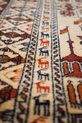A patterned carpet with intricate geometric designs and earthy tones, featuring detailed motifs and repetitive patterns. The texture appears soft, and the craftsmanship suggests a traditional or cultural style.