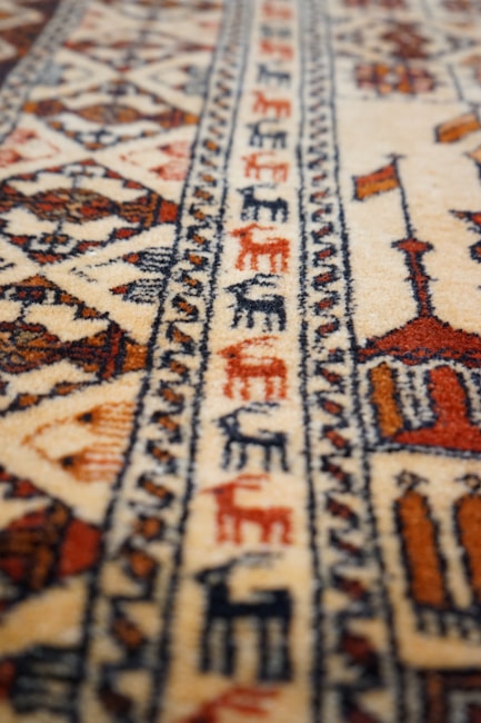 A patterned carpet with intricate geometric designs and earthy tones, featuring detailed motifs and repetitive patterns. The texture appears soft, and the craftsmanship suggests a traditional or cultural style.