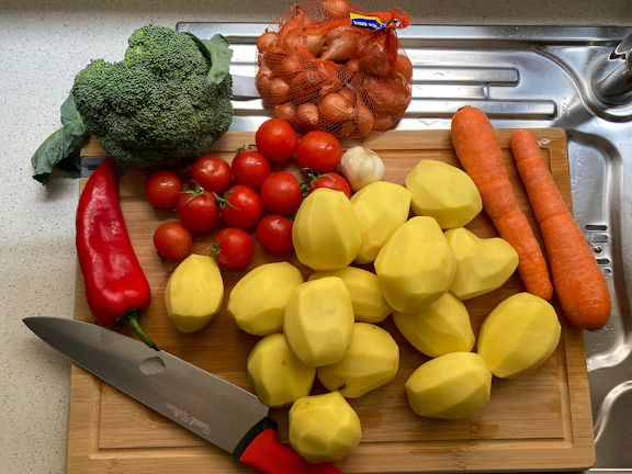 Close-up of a sleek stainless steel peeler resting on a wooden cutting board with fresh vegetables around.