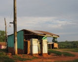 A rustic, small gas station with two vintage fuel pumps stands in a rural area. The building is constructed from weathered wood and painted in a faded green. The roof is corrugated metal, and there is a dirt road in front of the station. The surrounding area is lush with green grass and trees under a clear sky.