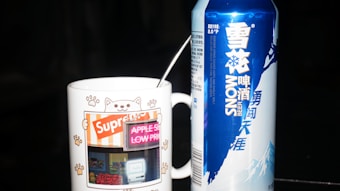 A white ceramic mug with cat paw prints and an illustration of a cat on it stands beside a tall blue and white can of Snow beer. The mug features graphics of a shop with a cat-themed design and the word 'Supreme'. A spoon is placed inside the mug.