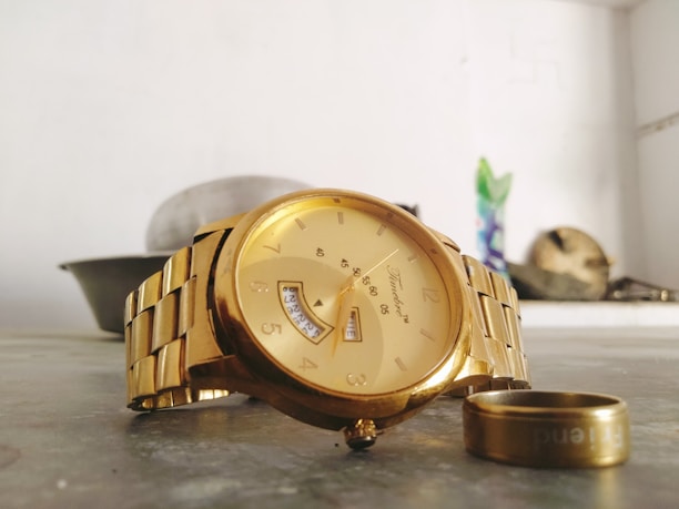 A gold-colored wristwatch with a metal band is prominently displayed on a surface. Next to the watch, there is a gold ring. In the background, a blurred setting with household items is visible, including a bowl and a decorative item, adding depth to the image.
