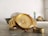 A gold-colored wristwatch with a metal band is prominently displayed on a surface. Next to the watch, there is a gold ring. In the background, a blurred setting with household items is visible, including a bowl and a decorative item, adding depth to the image.