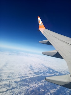 An airplane wing with a distinct orange and white logo is visible against a backdrop of clear blue sky and a layer of fluffy white clouds below.