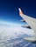 An airplane wing with a distinct orange and white logo is visible against a backdrop of clear blue sky and a layer of fluffy white clouds below.