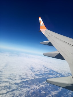 An airplane wing with a distinct orange and white logo is visible against a backdrop of clear blue sky and a layer of fluffy white clouds below.