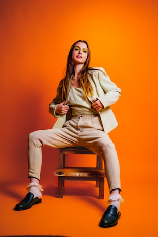 A person in a beige outfit sits confidently on a wooden stool against a vibrant orange background. The individual has long hair and wears colorful eye makeup, with a poised, assertive posture that adds a dynamic element to the composition.