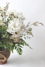 A bouquet of white flowers, likely daisies, arranged with green ferns and smaller white blooms. The bouquet is placed in a woven basket, creating a natural and rustic appearance.