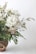 A bouquet of white flowers, likely daisies, arranged with green ferns and smaller white blooms. The bouquet is placed in a woven basket, creating a natural and rustic appearance.