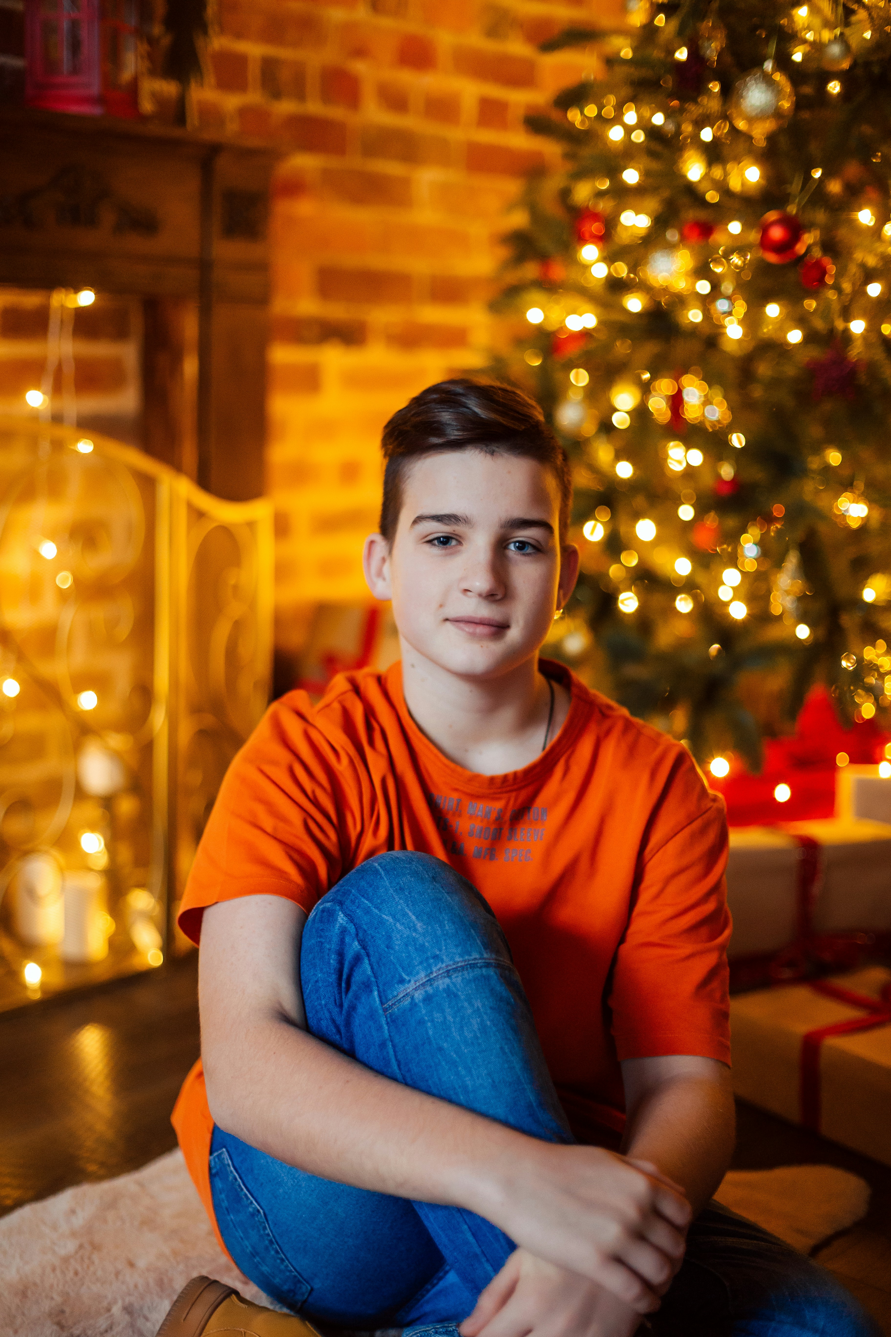 A smiling teenage boy sitting by a lit Christmas tree