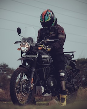 A person wearing a colorful helmet and black protective gear stands beside a classic Royal Enfield motorcycle in an outdoor setting. The landscape in the background is grassy with a cloudy sky, creating a moody atmosphere. The motorcycle is equipped with a windshield and has a rugged appearance, designed for adventure.