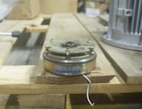 A mechanical clutch component with a metal casing and visible brand and model information is placed on a wooden pallet. The background includes industrial equipment, suggesting a workshop or manufacturing setting.