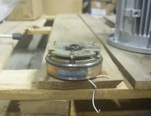 A mechanical clutch component with a metal casing and visible brand and model information is placed on a wooden pallet. The background includes industrial equipment, suggesting a workshop or manufacturing setting.