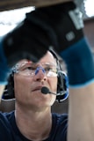 A person wearing safety goggles and a headset with a microphone is looking upwards. The individual is also wearing protective gloves and appears to be working or focusing on a task above them.