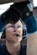 A person wearing safety goggles and a headset with a microphone is looking upwards. The individual is also wearing protective gloves and appears to be working or focusing on a task above them.