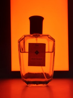 A transparent perfume bottle with dark edges and a black cap is set against a vibrant orange background. The label on the bottle reads 'Rayhan'. The lighting creates a silhouette effect around the bottle.