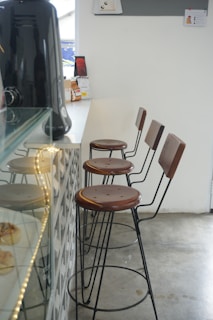 A modern cafe interior featuring a row of four high bar stools with metal frames and wooden seats. The stools are aligned along a counter with a coffee machine visible on the left. The flooring is concrete, and the walls are painted white, giving the space a minimalist aesthetic. On the counter, packaged goods and a small sign are placed near the window, which lets in natural light.