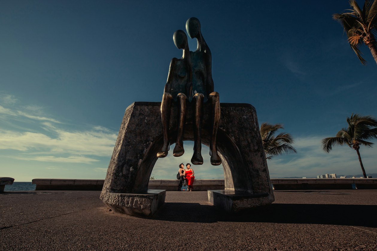 Two elongated bronze figures seated on a stone arch on the Puerto Vallarta Malecon at dusk