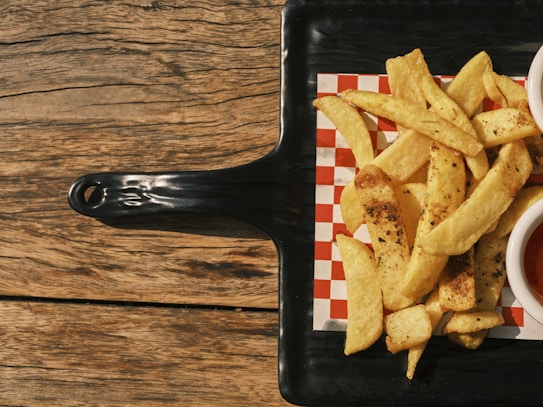A serving of golden-brown French fries seasoned with herbs rests on a square, black serving tray with a red and white checkered paper. The tray is placed on a rustic wooden table. A partial view of a cup with a red dipping sauce is visible beside the fries.