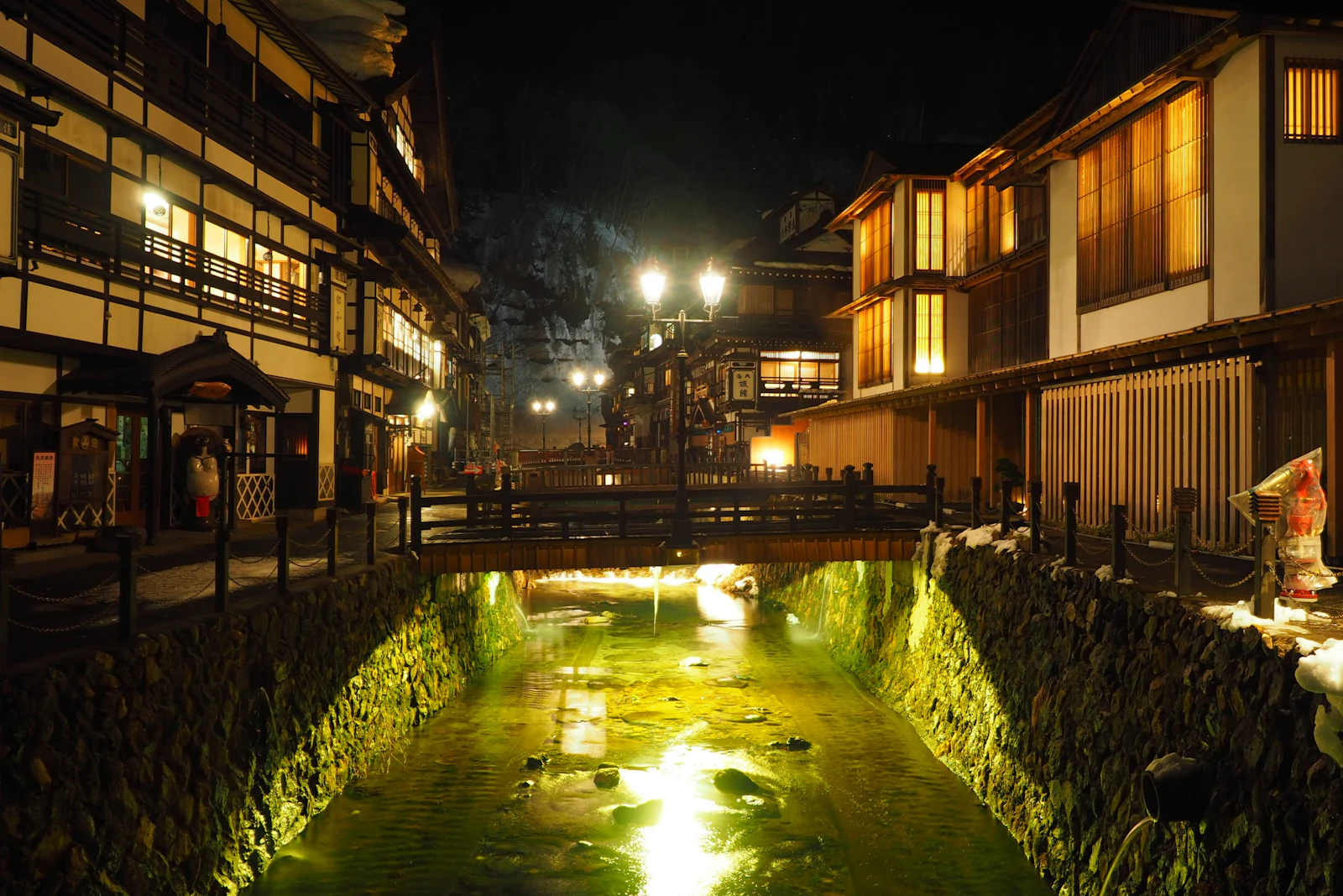 雪の残る冬の夜の銀山温泉。銀山川をはさんで木造旅館が並び、ガス燈と川のライトが灯っている