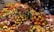 A vibrant array of fresh fruits displayed in a market setting, featuring a diverse selection including melons, bananas, pears, and mangoes. The fruits are arranged in a colorful and abundant manner, creating an inviting and lively atmosphere. The backdrop suggests a bustling market environment with various stalls in the background.