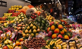 A vibrant array of fresh fruits displayed in a market setting, featuring a diverse selection including melons, bananas, pears, and mangoes. The fruits are arranged in a colorful and abundant manner, creating an inviting and lively atmosphere. The backdrop suggests a bustling market environment with various stalls in the background.