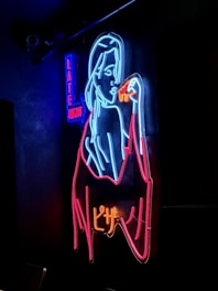 A neon sign featuring the outline of a woman holding a slice of pizza, depicted in vibrant colors. The words 'Late Night' can be seen in blue and red letters on the left side.