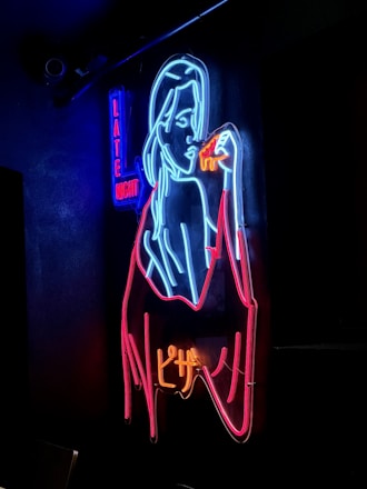 A neon sign featuring the outline of a woman holding a slice of pizza, depicted in vibrant colors. The words 'Late Night' can be seen in blue and red letters on the left side.