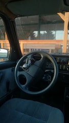 The interior of a car with a focus on the steering wheel, dashboard, and driver's seat. Through the windshield, a wooden structure and trees are visible. The steering wheel carries a logo, and keys are inserted in the ignition.