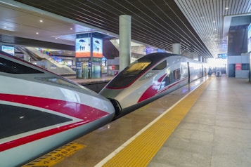 A modern high-speed train is stationed at a train platform inside a large, well-lit train station. The train features a sleek and aerodynamic design with a red, white, and gray color scheme. Overhead signs display information, and a few passengers can be seen near the platform edge. The station has an interior with pillars, digital screens, and overhead lights, adding a futuristic atmosphere.