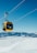 A bright yellow cable car suspended by thick cables, set against a backdrop of snow-covered mountains and a clear blue sky. The scene suggests a winter landscape with expansive views and crisp weather.