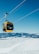 A bright yellow cable car suspended by thick cables, set against a backdrop of snow-covered mountains and a clear blue sky. The scene suggests a winter landscape with expansive views and crisp weather.