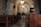 Inside a rustic stone church, there are wooden pews arranged in rows. The arching stone ceiling is illuminated by elegant chandeliers. A wooden cross is mounted on a stone wall at the front, enhancing the spiritual ambiance of the space.