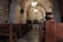 Inside a rustic stone church, there are wooden pews arranged in rows. The arching stone ceiling is illuminated by elegant chandeliers. A wooden cross is mounted on a stone wall at the front, enhancing the spiritual ambiance of the space.