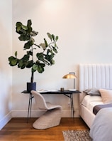 A cozy and modern bedroom corner with a small desk holding a potted plant. The desk features a sleek, minimalist design with a gold-colored lamp and an open book. A wicker chair complements the natural look. The room includes a plush bed with a light tufted headboard and soft bedding. The decor is calm and elegantly arranged, with wooden flooring adding warmth.