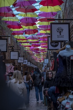 A bustling alleyway market is adorned with rows of colorful umbrellas hanging overhead. Shoppers, including a mannequin displaying clothes, move through the narrow space lined with stores. Signs promoting various brands and products are visible. The overall atmosphere is dynamic and lively, with a mixture of modern and vintage elements.