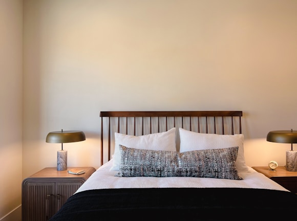 A neatly arranged bedroom with a wooden headboard against a pale wall, flanked by two wooden nightstands. Each nightstand holds a modern lamp with a metallic base and smooth, round shade. The bed is dressed in white linens with a textured design, complemented by a large, patterned throw pillow and a black blanket. A small clock and a book are placed on the nightstands.