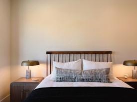 A neatly arranged bedroom with a wooden headboard against a pale wall, flanked by two wooden nightstands. Each nightstand holds a modern lamp with a metallic base and smooth, round shade. The bed is dressed in white linens with a textured design, complemented by a large, patterned throw pillow and a black blanket. A small clock and a book are placed on the nightstands.